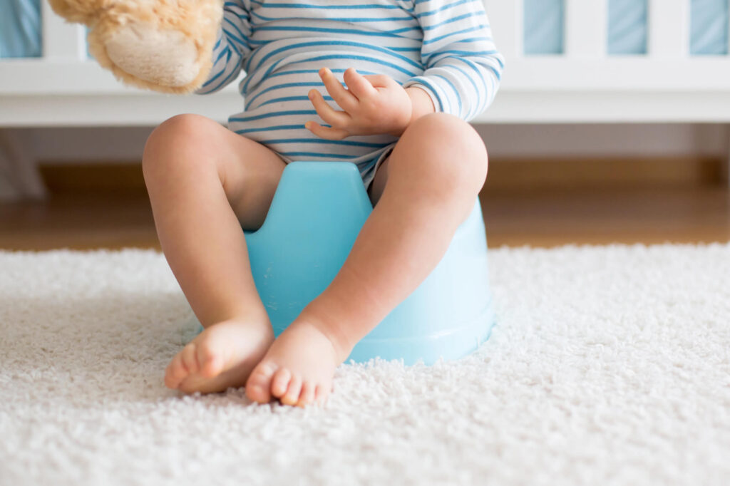 Toddler sitting on a blue potty seat holding a teddy bear during potty training